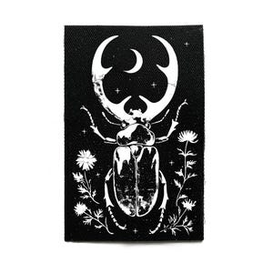 May include: A black rectangular patch with a white illustration of a beetle, a crescent moon, and floral elements. The beetle has large, curved horns. The design is detailed and artistic.