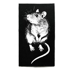 Puede incluir: Parche rectangular negro con una ilustración blanca de una rata. La rata se muestra sentada, con las patas juntas y una larga cola. El diseño es una representación artística detallada, ideal para coser en ropa o bolsos.