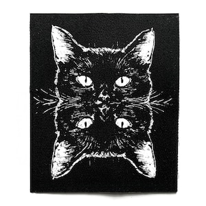 Peut inclure: Écusson noir et blanc représentant un visage de chat en miroir. Les yeux et les moustaches du chat sont blancs sur un fond noir. L'écusson est carré et en tissu texturé.