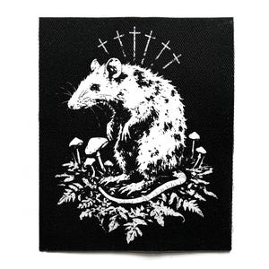 Puede incluir: Parche en blanco y negro con una rata estilizada rodeada de setas y follaje. Encima de la rata hay varios símbolos de cruz. El parche es cuadrado y está hecho de tela texturizada.