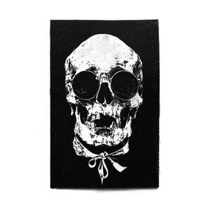 Könnte beinhalten: Schwarzer rechteckiger Aufnäher mit einem weißen Totenkopf, der eine runde Sonnenbrille und eine Fliege trägt. Der Schädel hat detaillierte Schattierungen, die ein strukturiertes Aussehen erzeugen. Das Design ist auf schwarzem Stoff gedruckt.