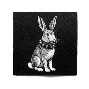 Könnte beinhalten: Schwarz-weißer Aufnäher mit einer detaillierten Illustration eines Kaninchens mit einem Nietenhalsband. Das Kaninchen sitzt aufrecht mit aufgestellten Ohren. Der Aufnäher hat eine quadratische Form und ist wahrscheinlich aus Stoff.
