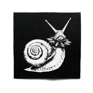 Könnte beinhalten: Ein quadratischer schwarzer Stoffaufnäher mit einer weißen Siebdruckillustration einer Schnecke. Die Schnecke ist mit einem detaillierten Spiralgehäuse dargestellt und trägt ein Halsband, das mit mehreren spitzen Metallstacheln besetzt ist.