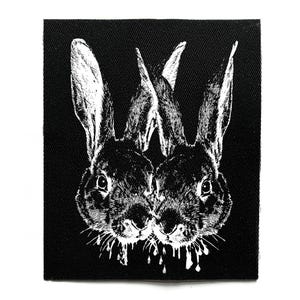 Pode incluir: Patch em preto e branco com uma ilustração gráfica de dois coelhos voltados um para o outro. Os coelhos têm orelhas longas e traços faciais detalhados. O design tem um estilo artístico desgastado, com detalhes gotejantes.