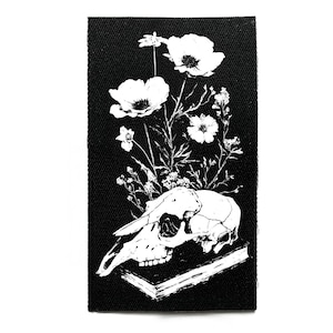 以下が含まれることがあります： 黒と白のパッチ。本の上に骸骨、白い花と葉が描かれています。デザインは詳細なイラストで、ゴシックまたはダークな美学です。長方形の形をしています。