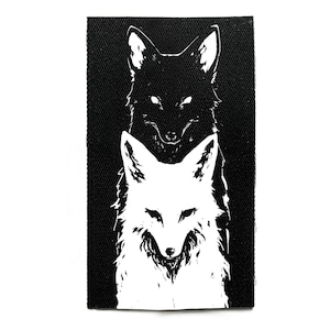 Könnte beinhalten: Schwarz-weißer Aufnäher mit zwei stilisierten Fuchsköpfen. Der obere Fuchs ist schwarz mit leuchtenden Augen, und der untere Fuchs ist weiß. Der Aufnäher hat einen strukturierten, dunklen Hintergrund.