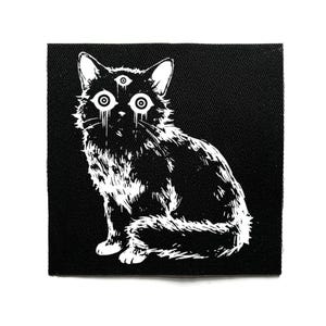 Pode incluir: Patch preto e branco com um gato estilizado de três olhos. O gato está sentado e tem uma aparência fofa. O design tem uma estética gótica ou alternativa.