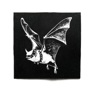 Op de afbeelding: Zwart vierkantige patch met een witte illustratie van een vleermuis in vlucht. De vleermuis draagt een spijkerhalsband. Het gedetailleerde ontwerp toont vleugel- en lichaamstexturen. Deze patch is waarschijnlijk voor kleding of accessoires.