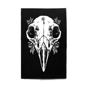 Peut inclure: Un écusson rectangulaire noir avec une illustration blanche d'un crâne d'oiseau et des détails botaniques. Le crâne a de grandes orbites et un long bec pointu. Le design est détaillé et de style gothique.