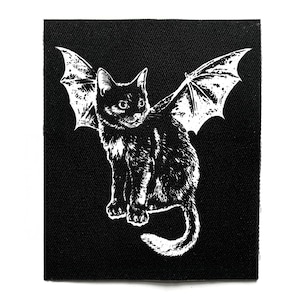 Puede incluir: Parche en blanco y negro con un gato con alas de murciélago. El gato está sentado con la cola enroscada. Las alas están extendidas detrás del gato. El parche está sobre un fondo negro.