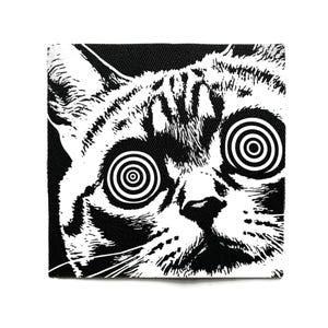 Peut inclure: Écusson carré noir et blanc représentant un visage de chat avec des yeux hypnotiques en spirale. La fourrure du chat est détaillée avec des rayures noires et blanches. Les yeux du chat sont le point central, avec des cercles concentriques créant un effet hypnotique.
