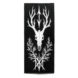 Puede incluir: Parche rectangular negro con un gráfico blanco de una calavera de ciervo con cuernos, una flecha y elementos botánicos. El diseño es simétrico y detallado, creando un contraste llamativo. El parche es probablemente para ropa o accesorios.