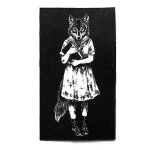 Może przedstawiać: Czarno-biała naszywka przedstawiająca postać z głową wilka w sukience marynarskiej, trzymającą książkę. Ilustracja ma estetykę vintage, ze szczegółami sierści i fałd sukienki. Naszywka jest prostokątna.