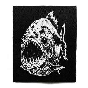 Könnte beinhalten: Ein rechteckiger schwarzer Stoffaufnäher mit einer kontrastreichen weißen Siebdruckillustration eines Piranhas. Der Fisch ist mit weit geöffnetem Maul dargestellt, das spitze Zähne zeigt, und weist ein detailliertes Schuppenmuster auf.