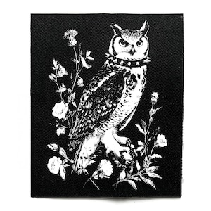 May include: Black and white patch featuring an owl with a studded collar, perched amidst floral elements. The owl is detailed with intricate feather patterns. The patch is square-shaped and made of a textured fabric.