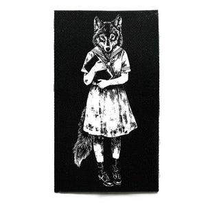 Op de afbeelding: Zwart-wit patch met een wolfskopfiguur in een matrozenjurk, die een boek vasthoudt. De illustratie heeft een vintage esthetiek, met gedetailleerde lijnen en een getextureerd uiterlijk. De patch is rechthoekig.