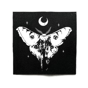 May include: Black square patch with a white moth illustration, a crescent moon, and four crosses. The moth has detailed wings and eye-like markings. The design is set against a black background, creating a striking contrast.