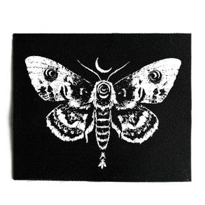 May include: A black square patch featuring a detailed white moth design. The moth has intricate wing patterns, a crescent moon above its head, and a stylized body. The patch is suitable for jackets, bags, or other fabric items.