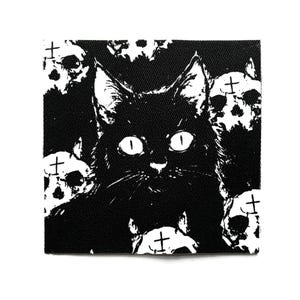 Könnte beinhalten: Quadratischer schwarz-weißer Aufnäher mit einer schwarzen Katze mit großen Augen, umgeben von weißen Totenköpfen. Das Design hat ein gotisches oder alternatives Thema.