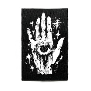 Può includere: Toppa rettangolare nera con un'illustrazione bianca di una mano aperta, un occhio nel palmo e stelle bianche. La toppa è realizzata in tessuto strutturato.