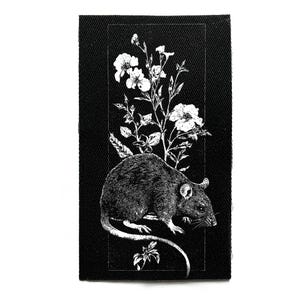 Puede incluir: Parche en blanco y negro con una ilustración detallada de una rata y flores blancas. El diseño está enmarcado en un rectángulo. La rata tiene una larga cola y está rodeada de delicado follaje.