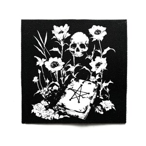 Könnte beinhalten: Schwarz-weißer Aufnäher mit einem gotischen Design. Es zeigt einen Schädel, Blumen und ein Buch mit einem Pentagramm. Die Illustration hat detaillierte florale Elemente. Der Aufnäher ist quadratisch und aus Stoff.
