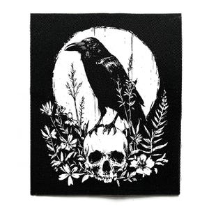 Può includere: Toppa in bianco e nero raffigurante un corvo appollaiato su un teschio, circondato da fiori e piante. Il corvo è in silhouette contro un cerchio bianco. La toppa ha uno sfondo nero, con dettagli dell'uccello e degli elementi.