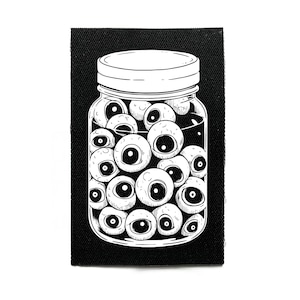 Könnte beinhalten: Schwarz-weißer Aufnäher mit einem Glas voller Augäpfel. Das Glas hat einen weißen Deckel und ist mit zahlreichen Augäpfeln mit schwarzen Pupillen gefüllt. Der Aufnäher ist rechteckig und vor schwarzem Hintergrund.