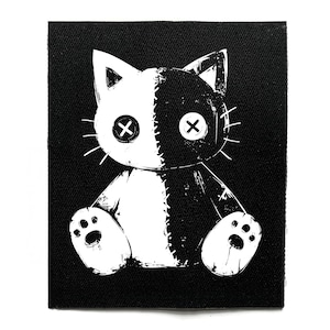 May include: Black and white patch featuring a split-faced cat design. One side is white, the other black, with stitched details and cross-shaped eyes. The cat is sitting, with paw prints visible.