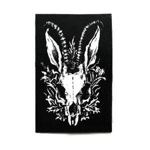 Może przedstawiać: Czarna, prostokątna naszywka z białą ilustracją czaszki kozy, dużymi rogami i elementami kwiatowymi. Gotycki design nadaje się do odzieży lub akcesoriów. Naszywka jest prostokątna.