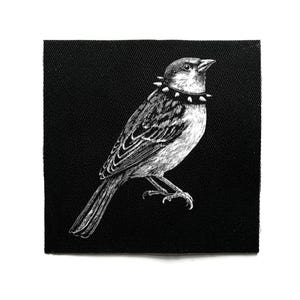以下が含まれることがあります： 黒と白のパッチ。スタッズカラーをつけた鳥の精巧なイラスト。鳥はとまり木に止まっており、羽や特徴が細かく描かれています。パッチは正方形で、黒い背景に配置されています。