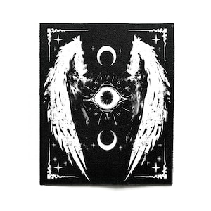Puede incluir: Parche en blanco y negro con una ilustración detallada. El diseño incluye un ojo central, lunas crecientes y alas estilizadas. El parche tiene un borde decorativo con estrellas.