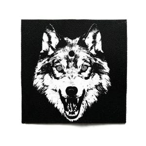 Può includere: Toppa in bianco e nero con un'illustrazione dettagliata della testa di un lupo. Il lupo ha la bocca aperta, mostrando i denti, e occhi a spirale. Un piccolo punto è sopra gli occhi. La toppa è quadrata.