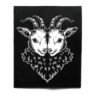 Può includere: Toppa quadrata nera con un'illustrazione bianca di due teste di capra rivolte l'una verso l'altra. Le capre hanno corna dettagliate e un'area del collo soffice, creando un design simmetrico. La toppa è probabilmente per abbigliamento o accessori.