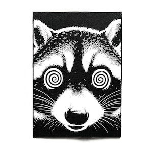 Peut inclure: Un écusson rectangulaire en tissu présentant une illustration graphique en noir et blanc à fort contraste d'un visage de raton laveur. Le raton laveur a des spirales blanches marquées à la place des yeux, un nez sombre et des moustaches.