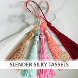 May include: A collection of six slender silky tassels in white, gold, light blue, pink, coral, and deep red. Each tassel features a matching coloured bead at the top and a thin cord loop for hanging. The text SLENDER SILKY TASSELS is visible.