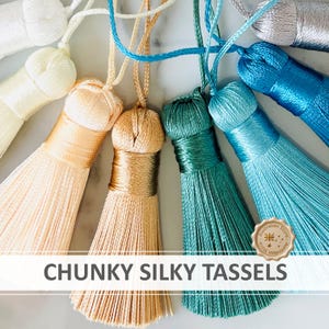 May include: A collection of chunky silky tassels in a fan arrangement, featuring colours of white, cream, peach, gold, teal, light blue, and silver. Each tassel has a braided hanging cord. The text CHUNKY SILKY TASSELS is visible at the bottom.