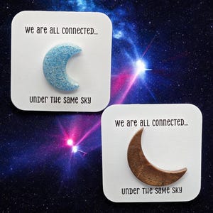 May include: Two white square cards with the text "we are all connected... under the same sky" printed on them. Each card features a crescent moon shape, one is blue with glitter and the other is brown.