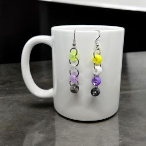 May include: A pair of dangle earrings with a silver chain and colorful beads. The beads are purple, white, yellow, and green. The earrings are hanging from a white mug.