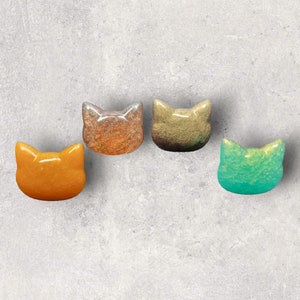 May include: Four ceramic cat head figurines in different colors. The cat heads are in a row, from left to right: orange, copper, gold, and teal.