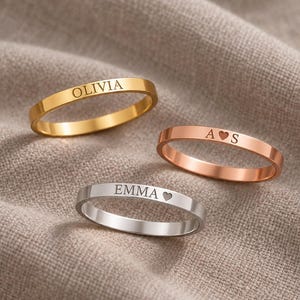 May include: Three thin metal rings in gold, rose gold, and silver tones. The gold ring is engraved with the name OLIVIA. The rose gold ring features the letters A and S separated by a heart. The silver ring is engraved with EMMA and a heart.