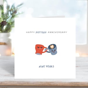 Pode incluir: Um cartão branco com o texto "HAPPY POTTERY ANNIVERSARY" e "NINE YEARS". O cartão apresenta duas canecas de desenho animado, uma vermelha e outra azul, de mãos dadas. A caneca azul tem um design em forma de coração.