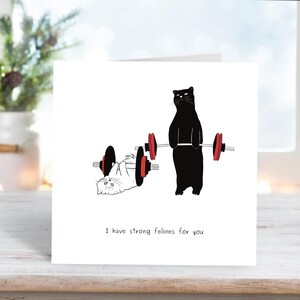 May include: A greeting card with a black cat lifting weights, a white cat lying on the ground, and the text "I have strong felines for you".