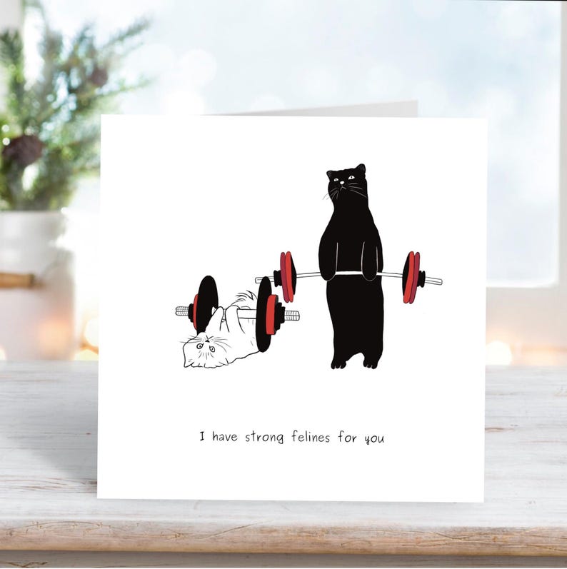 May include: A greeting card with a black cat lifting weights, a white cat lying on the ground, and the text "I have strong felines for you".
