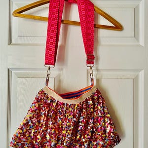 Peut inclure: Une jupe à sequins multicolores dotée d'une large ceinture dorée scintillante et d'une doublure intérieure rayée bleu et orange. La jupe est équipée d'une bandoulière réglable rose vif avec un motif répétitif d'étoiles et de carrés.