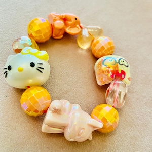 Könnte beinhalten: Ein Perlenarmband mit einem weißen Hello-Kitty-Anhänger, einer hellrosa Katzenfigur und einer kleinen gelben Figur mit Brille. Das Design umfasst mehrere runde, facettierte orangefarbene und gelbe Perlen mit einem reflektierenden, irisierenden Finish.