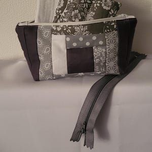Puede incluir: Un estuche rectangular de patchwork en tonos grises, blancos y negros. El estuche tiene una cremallera blanca y contiene un artículo a juego con estampado floral gris y blanco. Una cremallera gris está parcialmente desabrochada.