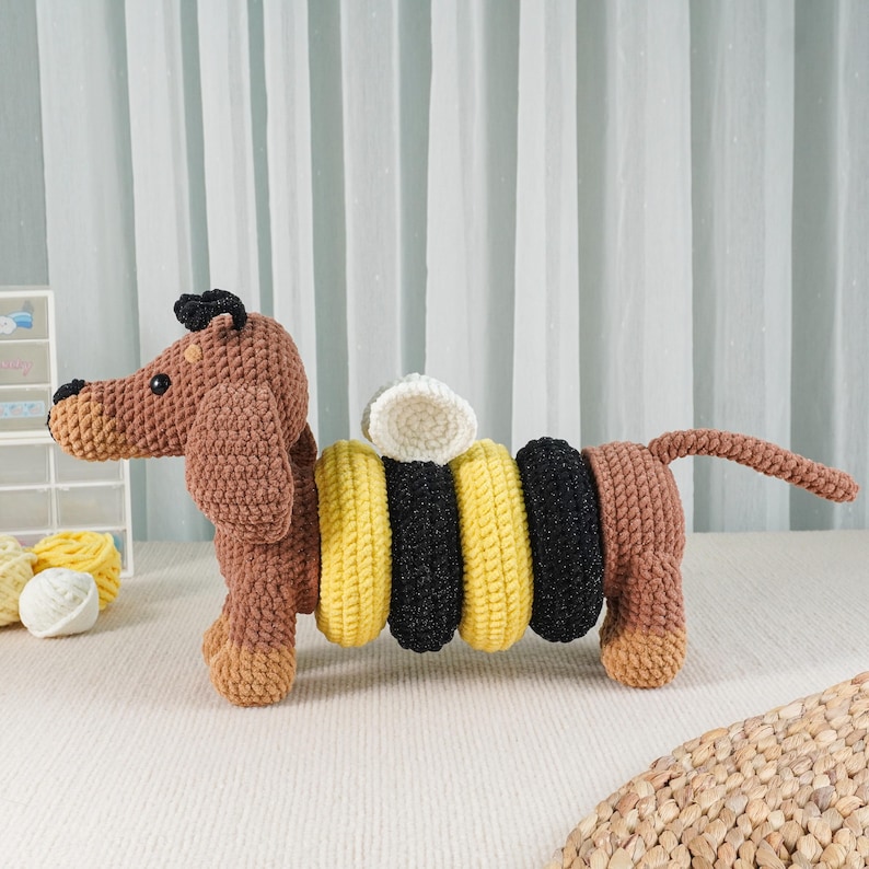 May include: A crocheted dachshund dog toy in shades of brown, yellow, black, and white. The toy has a segmented body with yellow and black rings, a white ring on top, and a small tail. The dog is standing on a white surface.