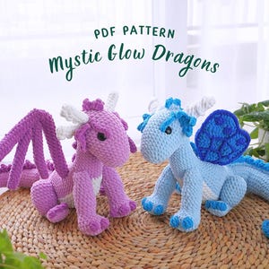May include: PDF pattern for crocheting two glow-in-the-dark dragons. One dragon is purple with white accents and the other is blue with white accents. Both dragons have wings and are sitting on a woven surface.