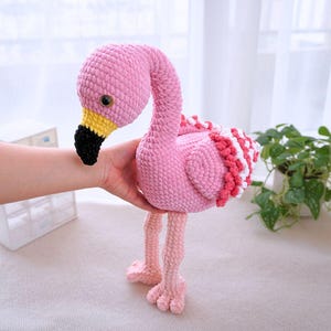 May include: A handmade pink crochet flamingo with a long, curved neck and a black beak. The flamingo has yellow accents on its beak and a ruffled design on its back with red and white details. The flamingo's legs and feet are a lighter shade of pink.